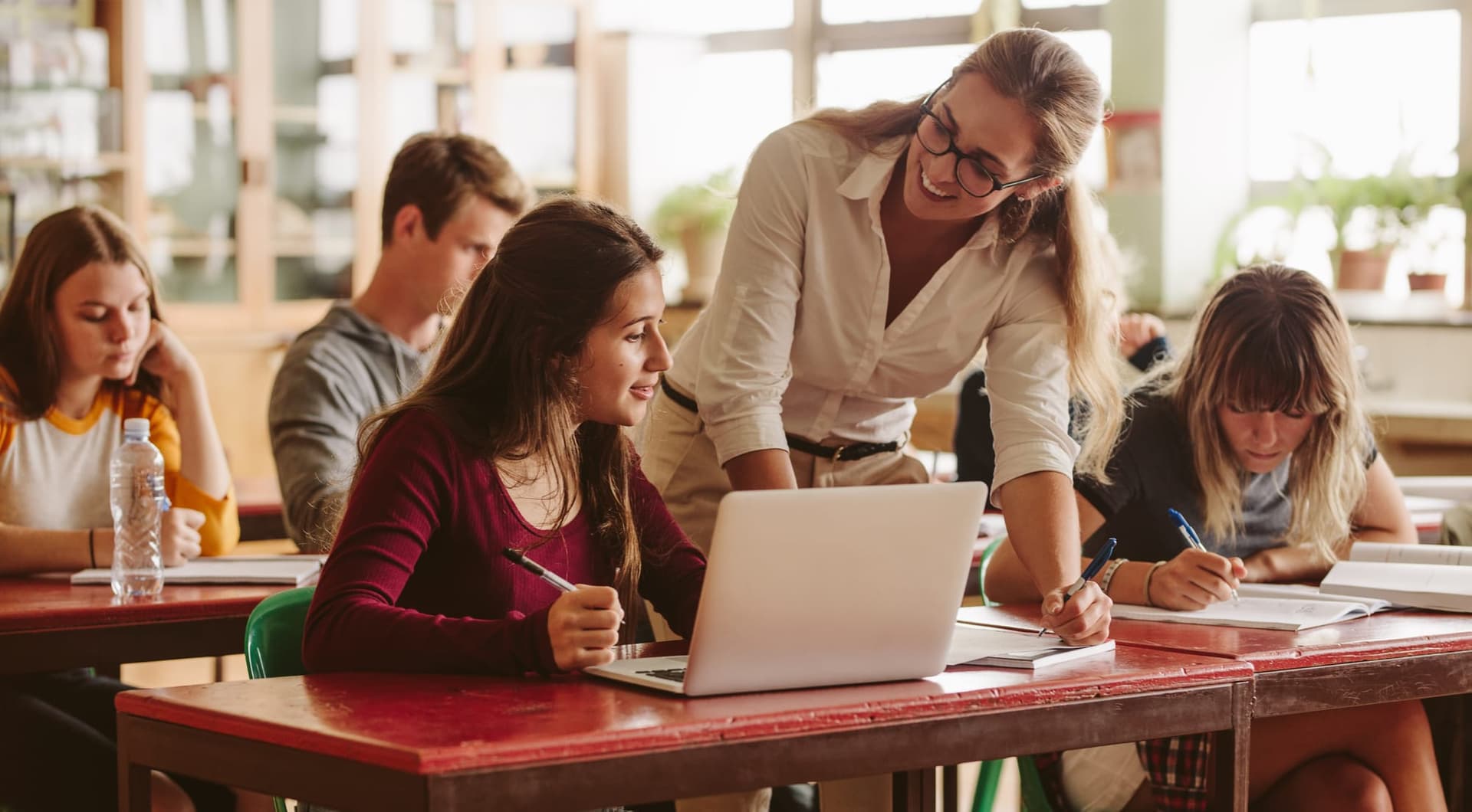 A smiling teacher leans over a high-school student's desk to help with work on a laptop while classmates study nearby